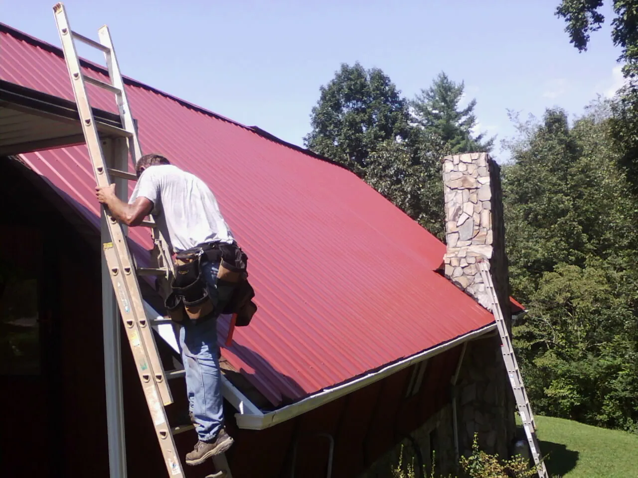 Roofer on a ladder beside a red metal roof and stone chimney — Soffit & Fascia Repair by Roofing Brothers in South Sioux City